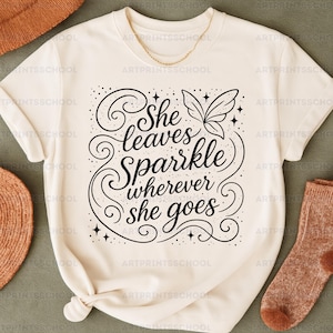 Pode incluir: Camiseta creme com a frase "She leaves Sparkle wherever she goes" em fonte preta decorativa. O design inclui redemoinhos, estrelas e uma borboleta. Um colar dourado e uma meia marrom também são visíveis.