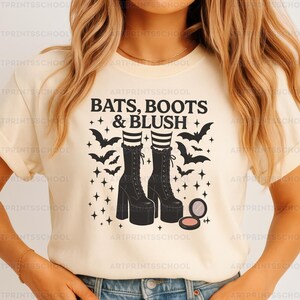 Puede incluir: Camiseta color crema con un diseño gráfico. El diseño presenta el texto "BATS, BOOTS & BLUSH", botas negras, murciélagos, estrellas y un estuche de maquillaje. Un diseño divertido y espeluznante para Halloween.