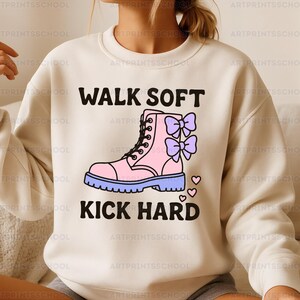 May include: Cream-colored sweatshirt with the text "WALK SOFT KICK HARD" above and below a pink boot graphic with purple bows and hearts. The boot has a blue sole and black laces. A fun, casual design.
