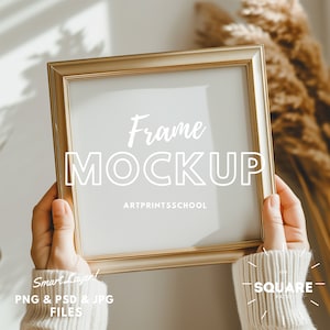 Può includere: Una cornice dorata con sfondo bianco e il testo "Frame MOCKUP ARTPRINTSSCHOOL" è tenuta da una persona che indossa un maglione bianco. La cornice è quadrata e ha un design semplice.