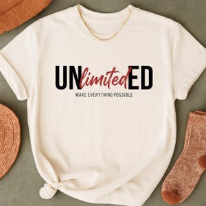 May include: Cream-colored t-shirt with the word "UNlimited" in black and red lettering. The phrase "MAKE EVERYTHING POSSIBLE" is printed below the main text. A gold chain necklace is visible at the neckline.