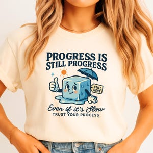 May include: Cream-colored t-shirt with the text "Progress is Still Progress" and "Even if it's Slow, Trust Your Process." The graphic features a smiling ice cube with an umbrella, giving a thumbs up.