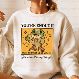 以下が含まれることがあります： クリーム色のスウェットシャツ。フード付きのパーカーと赤いスニーカーを履いたカエルのイラストが描かれ、親指を立てています。「YOU'RE ENOUGH」、「NEVER FORGET YOUR WORTH」、「You Are Already Magic」の文字があります。