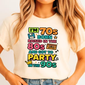 May include: Cream-colored t-shirt with a colorful graphic design. The design features the text "I'M 70s BORN RAISED IN THE 80s AND GOT TO PARTY IN THE 90s" with retro icons like a floppy disk and cassette tape.