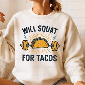 Pode incluir: Camisola creme com o texto "WILL SQUAT FOR TACOS" em preto. Um taco de desenho animado está centrado, com um design de haltere. O taco é amarelo com detalhes verdes, castanhos e laranja. Raios e raios de sol também estão incluídos.