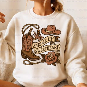 Puede incluir: Sudadera color crema con un gráfico de temática western. El diseño incluye una bota de vaquero, sombrero, herradura, cuerda y una pancarta con el texto "Saddle Up Sweetheart". También se incluye una rosa.