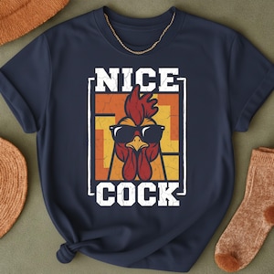 May include: Navy blue t-shirt featuring a rooster wearing sunglasses with the text "NICE COCK" in white. The rooster graphic is set against a square of orange and yellow. A gold chain is visible at the neckline.