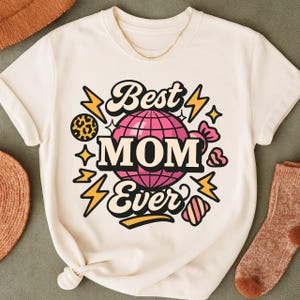 May include: Cream-coloured t-shirt featuring the text "Best MOM Ever" in a retro style. The design incorporates a pink globe, lightning bolts, and other decorative elements. A gold chain is visible at the neckline.