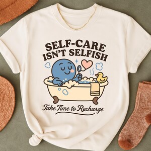 Puede incluir: Camiseta color crema con el texto "SELF-CARE ISN'T SELFISH" y "Take Time to Recharge". El gráfico presenta un pulpo azul en una bañera con burbujas, un corazón y un patito de goma. Se ve un collar dorado.