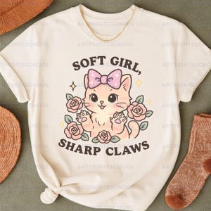 May include: Cream t-shirt with a cartoon cat design, pink bow, and roses. The text reads "SOFT GIRL, SHARP CLAWS". A gold chain is visible at the top. The design is printed on a white t-shirt.