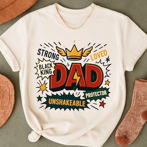 Pode incluir: Camiseta creme com um design gráfico. O design inclui a palavra "DAD" em letras vermelhas com um raio amarelo. Contém as palavras "STRONG", "BLACK KING", "LOVED", "PROTECTOR" e "UNSHAKEABLE". Uma coroa dourada com asas.