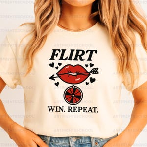 May include: Cream-colored t-shirt featuring the word "FLIRT" above a red lip pierced by an arrow, surrounded by hearts. Below, a roulette wheel graphic and the words "WIN. REPEAT." create a playful design. A casual, fun tee.