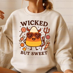 Puede incluir: Sudadera de color crema con un gráfico temático de Halloween. El diseño presenta un personaje de caramelo de maíz con gafas de sol en forma de corazón, cuernos de diablo y un tridente, junto con las palabras "WICKED BUT SWEET". Varios caramelos rodean el caramelo de maíz.
