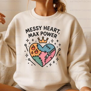 Puede incluir: Sudadera color crema con el texto "MESSY HEART, MAX POWER" sobre un diseño de corazón colorido. El corazón está parcheado con varios colores y presenta una cremallera, llamas y una corona.