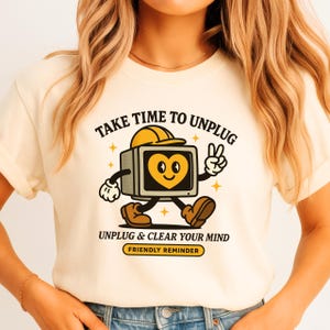 May include: Cream-colored t-shirt featuring a retro cartoon television character with a heart-shaped screen, wearing a hard hat and giving a peace sign. The shirt has the text "TAKE TIME TO UNPLUG" and "UNPLUG & CLEAR YOUR MIND".