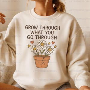 Puede incluir: Sudadera color crema con el texto negro "GROW THROUGH WHAT YOU GO THROUGH". Debajo, un gráfico de margaritas en una maceta marrón, con corazones y estrellas. La sudadera es de estilo casual.
