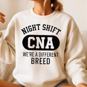 May include: Cream-colored sweatshirt with the text "NIGHT SHIFT CNA WE'RE A DIFFERENT BREED" in black. The word "CNA" is in a black oval. The sweatshirt is a crew neck style.