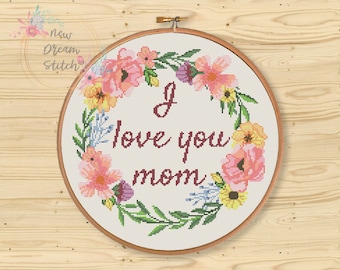 Puede incluir: Un aro de bordado blanco con un diseño de corona floral y el texto "I love you mom" en letras cursivas marrón.