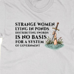 May include: A light gray t-shirt with the text "STRANGE WOMEN LYING IN PONDS DISTRIBUTING SWORDS IS NO BASIS FOR A SYSTEM OF GOVERNMENT." A cartoon shows a hand holding a sword emerging from water.