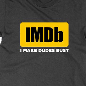 May include: Dark grey t-shirt featuring a yellow rectangle with the black letters "IMDb". Beneath, the white text reads "I MAKE DUDES BUST". The shirt has a crew neck design.