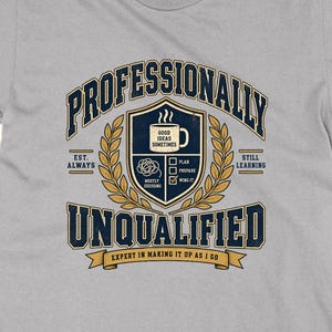 Könnte beinhalten: Ein hellgraues Kurzarm-T-Shirt mit einer dunkelblauen und goldenen Grafik. Das Design zeigt ein Wappen mit einer Kaffeetasse, einer Checkliste und dem Text: PROFESSIONALLY UNQUALIFIED, EST. ALWAYS, STILL LEARNING, EXPERT IN MAKING IT UP AS I GO.
