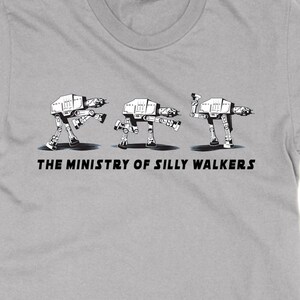 May include: A light gray t-shirt featuring a humorous graphic of three AT-AT walkers in various silly poses. The text "THE MINISTRY OF SILLY WALKERS" is printed below the graphic in black. The shirt is made of a soft material.