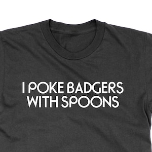 Peut inclure: Un t-shirt gris foncé avec le texte blanc "I POKE BADGERS WITH SPOONS". Ce t-shirt à manches courtes a un col rond. Le texte est centré sur le devant du t-shirt et est fait d'une matière douce.