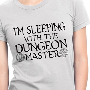 Op de afbeelding: Een wit T-shirt met zwarte tekst die "Ik slaap met de Dungeon Master" luidt, met een cirkelvormig ontwerp aan beide kanten van de tekst.