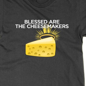 May include: Dark gray t-shirt with the text "BLESSED ARE THE CHEESEMAKERS" above a cartoon wedge of cheese with a halo. The cheese is yellow with several holes. The halo is gold with sun rays.