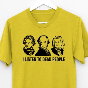 May include: Mustard yellow t-shirt with a black graphic of three historical figures and the text "I LISTEN TO DEAD PEOPLE". The shirt is hanging on a white hanger against a white background.