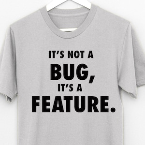 Könnte beinhalten: Ein graues T-Shirt mit schwarzem Text, der "Es ist kein Fehler, es ist eine Funktion." lautet.