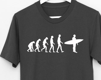 Puede incluir: Camiseta gris oscuro con un gráfico blanco que representa la evolución humana, culminando con un surfista con una tabla de surf. La camiseta está colgada en una percha blanca sobre un fondo blanco. Un diseño divertido para surfistas.