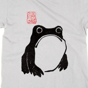 May include: A pale grey t-shirt with a black frog design and a large white belly. The frog has large eyes and a red Japanese stamp above it. The shirt is made of a soft material.
