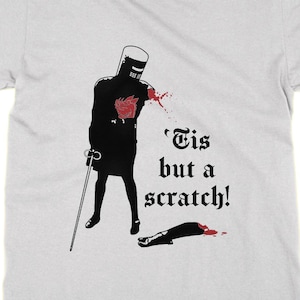 May include: Light gray t-shirt featuring a black illustration of a knight with a red wound and the text "'Tis but a scratch!". The knight is holding a cane. The design is centered on the shirt.