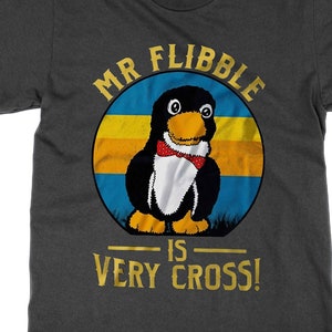 Op de afbeelding: Een donkergrijs T-shirt met een gouden afbeelding van een pinguïn met een strik. De afbeelding is omgeven door een cirkel met de tekst "Mr Flibble is Very Cross!" in goud.