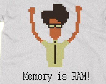 May include: A white t-shirt with a pixelated image of a person with brown hair and glasses, raising their arms in the air. The text "Memory is RAM!" is printed below the image.