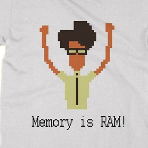 May include: A white t-shirt featuring a pixelated image of a person with brown hair and glasses, raising their arms in the air. The text "Memory is RAM!" is printed below the image.