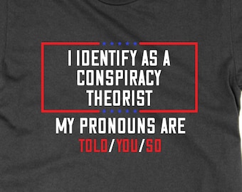 May include: A dark grey t-shirt with a red box and white text that reads "I identify as a conspiracy theorist. My pronouns are told/you/so."