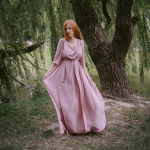 May include: A long, flowing dress in a dusty rose hue, featuring a draped neckline and a fitted waist. The dress has long sleeves and a full skirt. The woman in the dress has long red hair and is standing in a grassy area near a large tree.