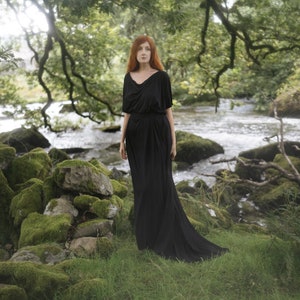 May include: A woman with long, red hair stands in a woodland setting, wearing a long, black dress. She is standing near a stream and rocks covered in moss.