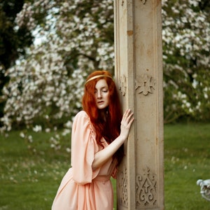 May include: A woman with long, red hair wearing a peach-colored dress stands next to a stone pillar with intricate carvings. The pillar is in a garden setting with green grass and white flowers.