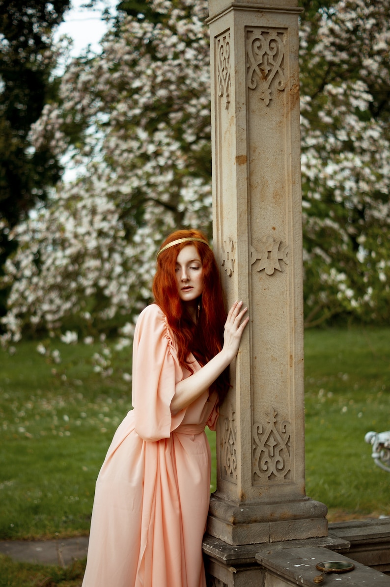 May include: A woman with long, red hair wearing a peach-colored dress stands next to a stone pillar with intricate carvings. The pillar is in a garden setting with green grass and white flowers.