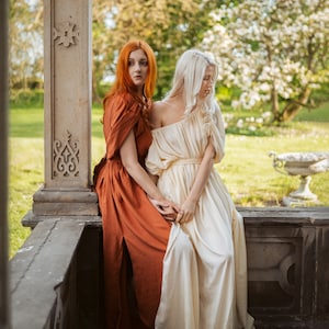 Puede incluir: Dos mujeres con vestidos largos, una con un vestido naranja &oacute;xido y la otra con un vestido blanco, est&aacute;n sentadas en un borde de piedra. La mujer con el vestido naranja mira hacia la izquierda, mientras que la mujer con el vestido blanco mira hacia abajo. El fondo es un exuberante jard&iacute;n verde con un pilar de piedra.