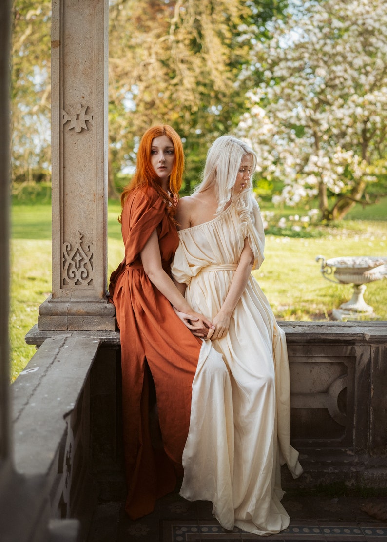 Puede incluir: Dos mujeres con vestidos largos, una con un vestido naranja &oacute;xido y la otra con un vestido blanco, est&aacute;n sentadas en un borde de piedra. La mujer con el vestido naranja mira hacia la izquierda, mientras que la mujer con el vestido blanco mira hacia abajo. El fondo es un exuberante jard&iacute;n verde con un pilar de piedra.