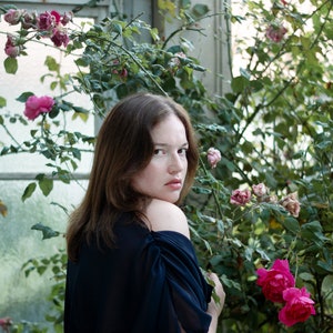 May include: A woman with long brown hair wearing a dark blue off-the-shoulder dress stands in front of a white wall with a rose bush growing in front of it. The woman is looking over her shoulder at the camera.