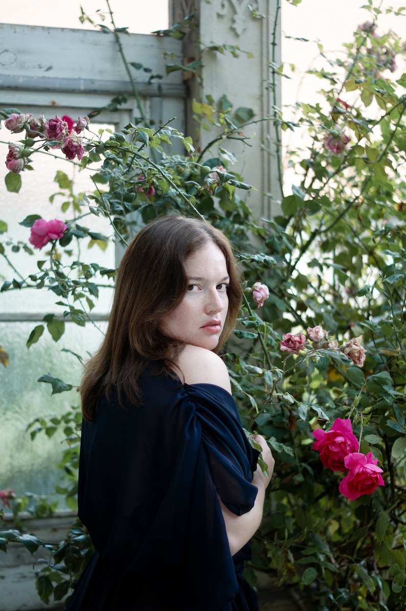 May include: A woman with long brown hair wearing a dark blue off-the-shoulder dress stands in front of a white wall with a rose bush growing in front of it. The woman is looking over her shoulder at the camera.