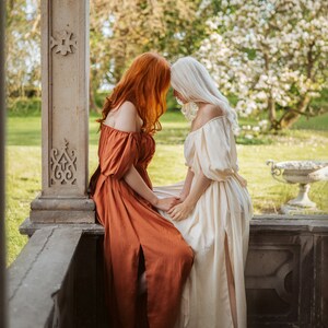 Puede incluir: Dos personas con vestidos largos, uno de color naranja &oacute;xido y el otro de color crema, est&aacute;n sentadas en un borde de piedra. Se miran de frente con las cabezas toc&aacute;ndose.