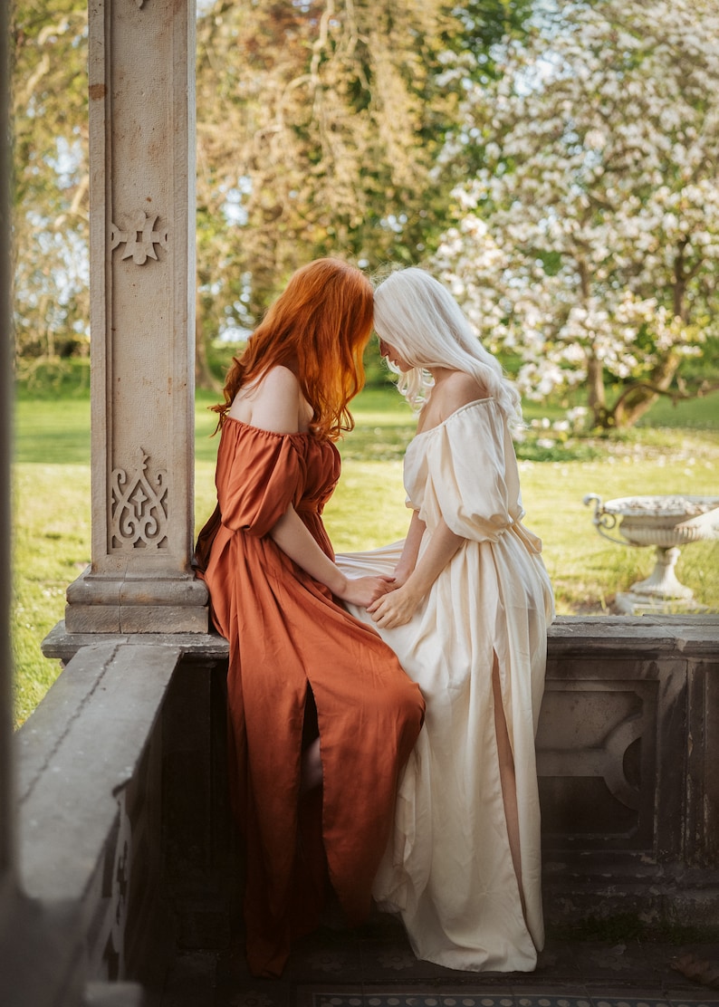 Puede incluir: Dos personas con vestidos largos, uno de color naranja &oacute;xido y el otro de color crema, est&aacute;n sentadas en un borde de piedra. Se miran de frente con las cabezas toc&aacute;ndose.