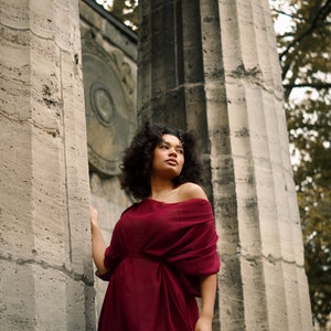 May include: A woman with curly hair wearing a burgundy off-the-shoulder dress stands between two stone pillars.
