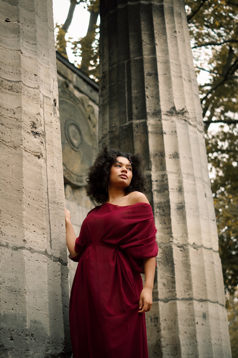 May include: A woman with curly hair wearing a burgundy off-the-shoulder dress stands between two stone pillars.